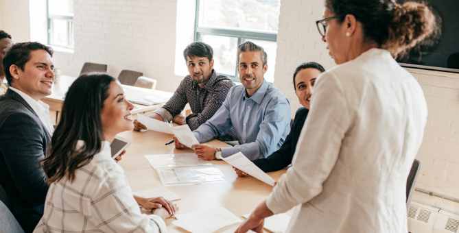 A group of people sitting around a conference table, holding papers and looking at a person standing and speaking.