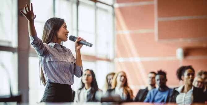 A woman speaking into a microphone with her hand raised, addressing an audience seated in a conference room.