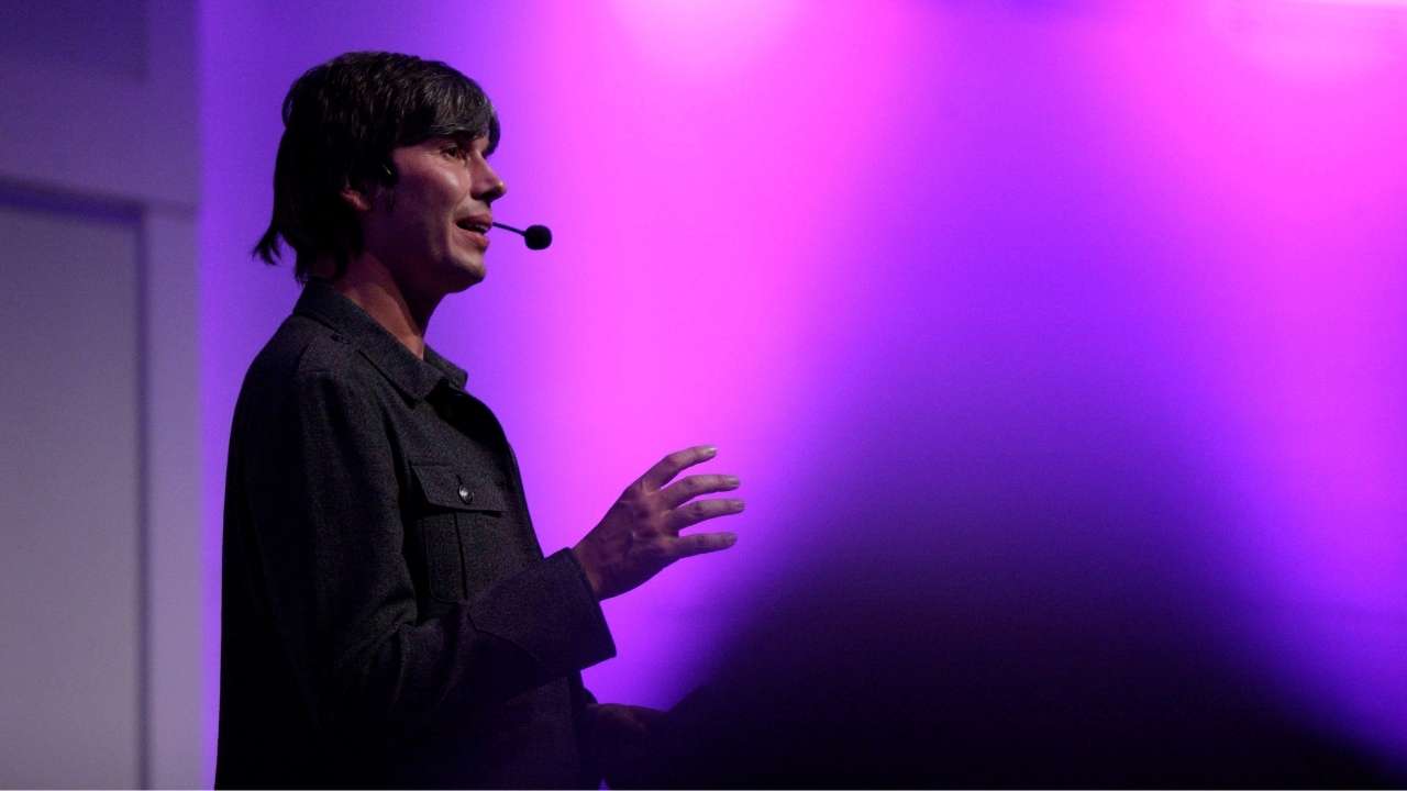 Professor Brian Cox is speaking confidently, wearing a headset microphone.