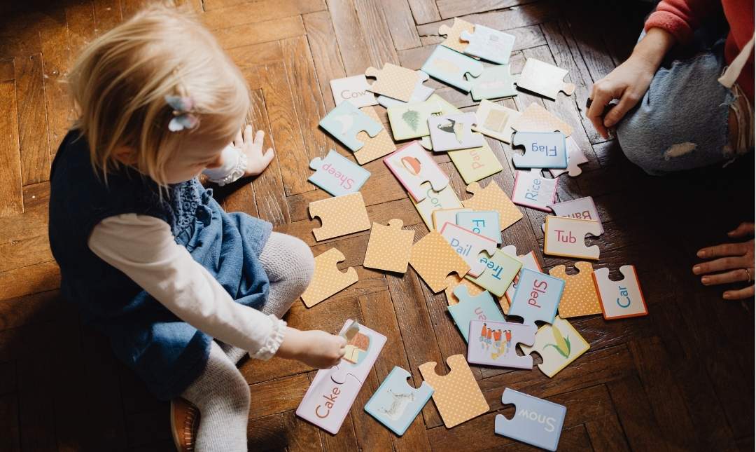 A young child sits on a wooden floor surrounded by scattered puzzle pieces with letters and pictures